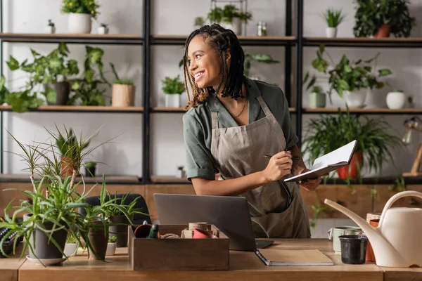 Cheerful african american florist with notebook looking away near laptop and potted plants in flower shop — Stock Photo