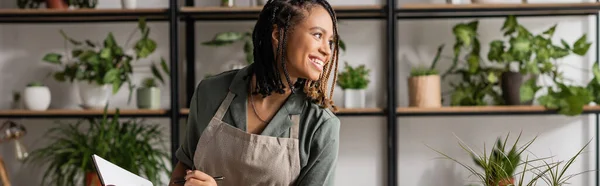 Cheerful african american florist holding notebook and looking away near rack with plants on blurred background, banner — Stock Photo