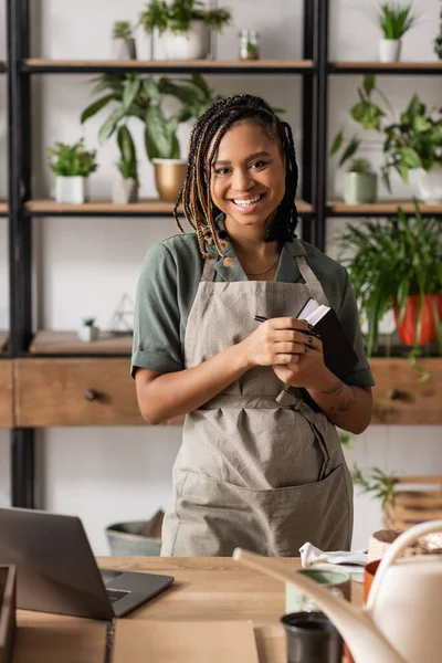 Trendy african american woman with copybook looking at camera near laptop and potted plants on blurred background — Stock Photo