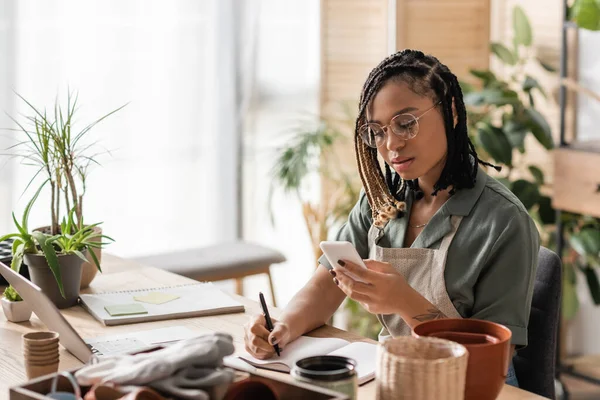 Young african american florist in eyeglasses looking at smartphone and writing order in notebook in flower shop — Stock Photo