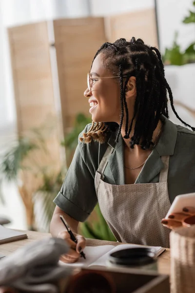 Smiling african american florist looking away while holding smartphone and writing order in flower shop on blurred foreground — Stock Photo