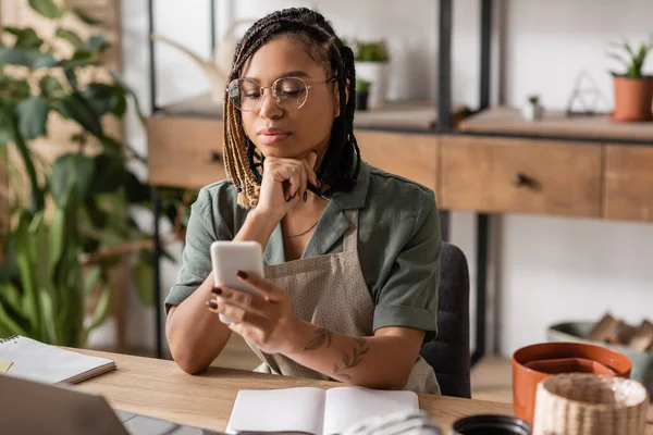 Thoughtful african american florist in eyeglasses looking at cellphone near blank notebook in flower shop — Stock Photo