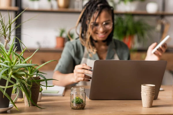 Sfocato fiorista afroamericano che lavora al computer portatile vicino a piante in vaso verdi nel negozio di fiori — Foto stock