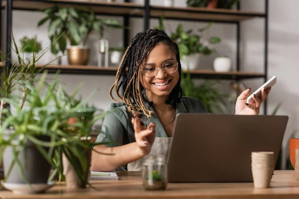 Happy african american florist holding smartphone and gesturing during video call on laptop near blurred plants — Stock Photo