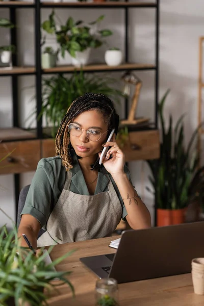 Fiorista afroamericano in grembiule e occhiali parlare sul cellulare vicino laptop e piante sfocate in negozio di fiori — Foto stock