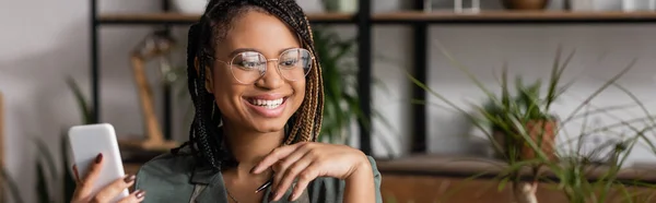 Fröhliche afrikanisch-amerikanische Floristin mit trendiger Frisur und Brille mit Handy im Blumenladen, Banner — Stockfoto