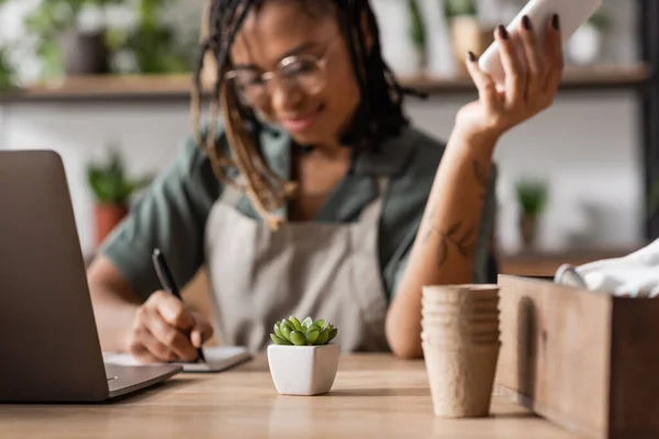Selective focus of small potted plant near laptop and african american florist writing order on blurred background — Stock Photo