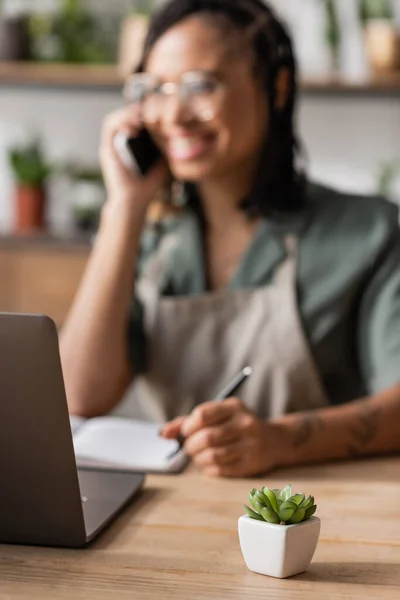 Selective focus of green potted plant near computer and african american florist talking on smartphone on blurred background — Stock Photo