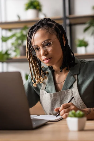Focused african american florist in eyeglasses looking at laptop and writing order in flower shop — Stock Photo