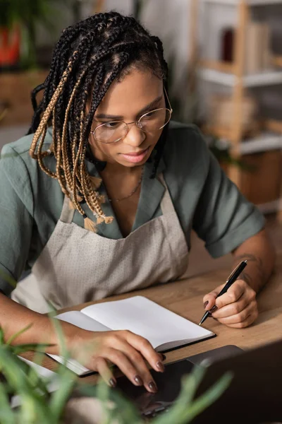 Африканский американский флорист с дредами с помощью ноутбука, держа в руках ручку рядом с пустым блокнотом в цветочном магазине — стоковое фото