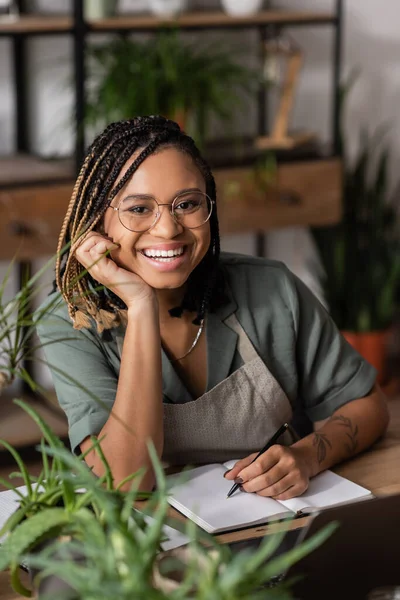Happy african american florist with dreadlocks and eyeglasses looking at camera near empty copybook in flower shop — Stock Photo
