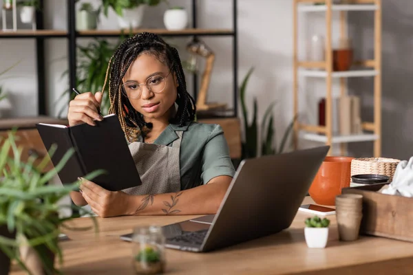 Elegante fiorista africano americano che tiene notebook mentre seduto sul posto di lavoro vicino al computer portatile in negozio di fiori — Foto stock
