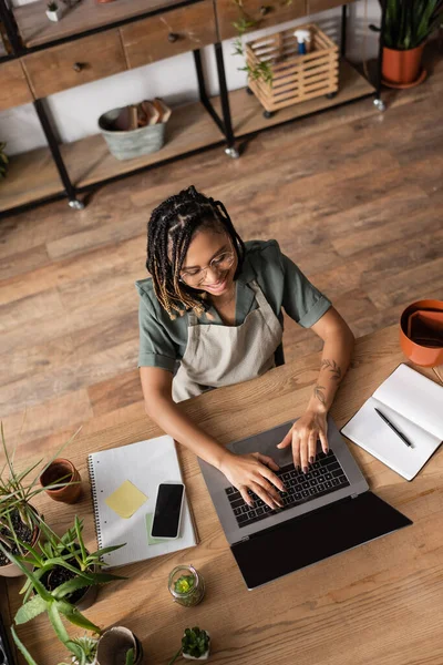 Top view of smiling african american florist typing on laptop near flowerpots and copybooks on desk in shop — Stock Photo