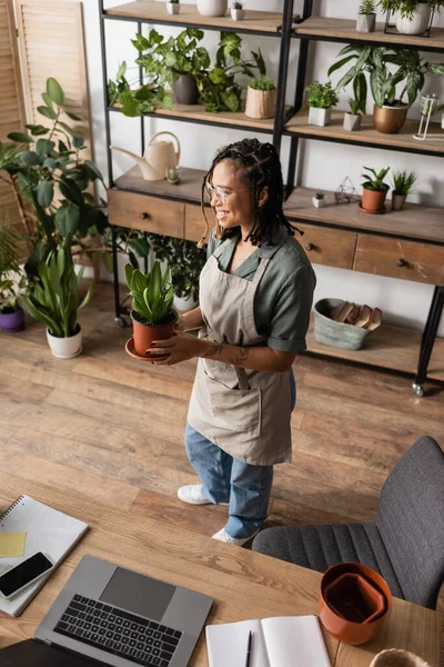 Vista ad alto angolo di felice fiorista afroamericano che tiene pianta in vaso vicino alla scrivania con dispositivi nel negozio di fiori — Foto stock