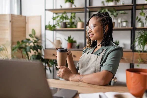 Happy african american florist in eyeglasses and apron sitting with paper cup near blurred laptop in flower shop — Stock Photo