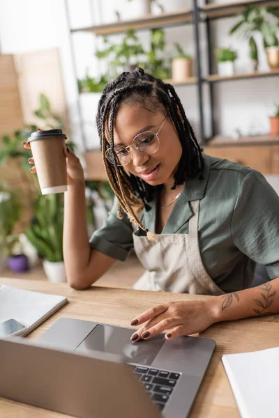 Stylish african american florist with coffee to go using laptop on desk in flower shop — Stock Photo