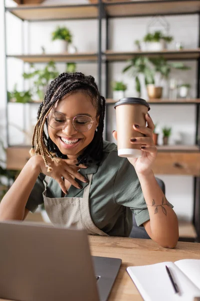 Fleuriste afro-américain à la mode avec café pour aller sourire près d'un ordinateur portable lors d'un appel vidéo dans un magasin de fleurs — Photo de stock