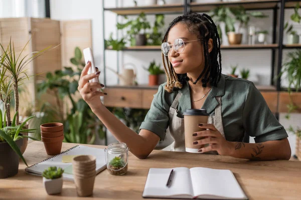 Smiling african american florist with paper cup looking at cellphone near empty notebook and flowerpots in shop — Stock Photo