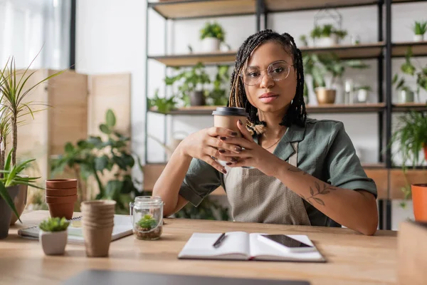African american florist with braids and eyeglasses holding paper cup and looking at camera near cellphone in flower shop — Stock Photo