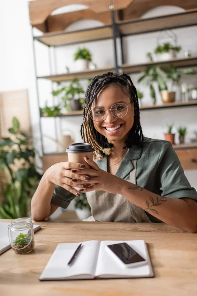 Happy african american florist with coffee to go looking at camera near smartphone and notebook on desk in shop — Stock Photo