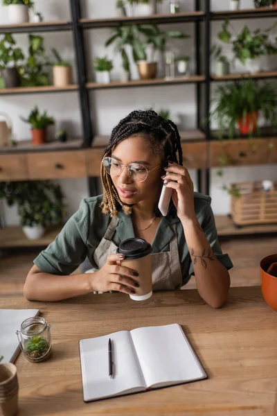 Young african american florist in apron and eyeglasses holding takeaway drink and talking on smartphone in flower shop — Stock Photo