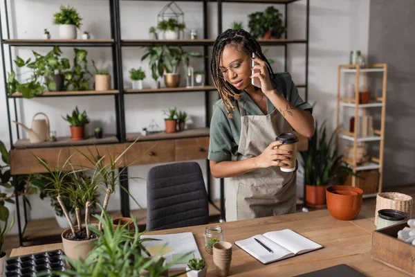 Floristería afroamericana con taza de papel hablando en el teléfono móvil y mirando el cuaderno en blanco cerca de plantas en maceta - foto de stock