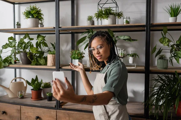 Jeune fleuriste afro-américain avec téléphone portable pointant vers un assortiment de plantes dans un magasin de fleurs — Photo de stock