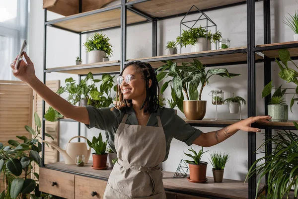 Floristería afroamericana sonriente con teléfono móvil de pie cerca de rack con plantas en maceta durante el pedido en línea - foto de stock