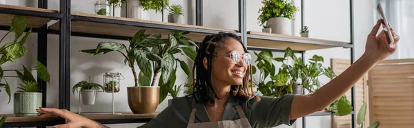 Smiling african american florist in eyeglasses showing assortment of potted plants during video call on smartphone, banner — Stock Photo