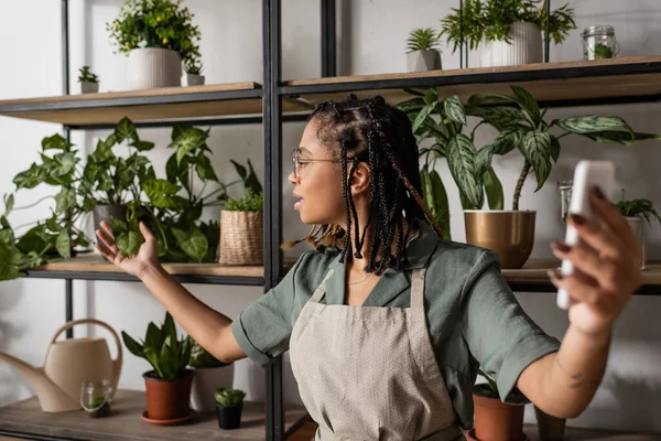 African american florist in apron holding smartphone and touching leaves of green plant during online trade — Stock Photo