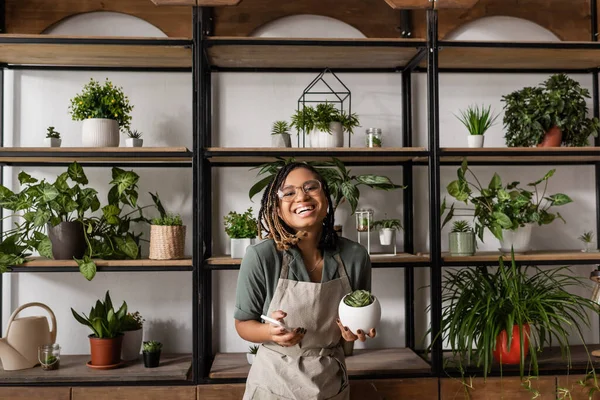 Happy african american florist with mobile phone looking at camera near rack with different potted plants — Stock Photo