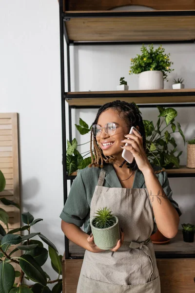 Souriant afro-américain fleuriste dans les lunettes et tablier tenant plante en pot et parler sur téléphone portable dans le magasin de fleurs — Photo de stock