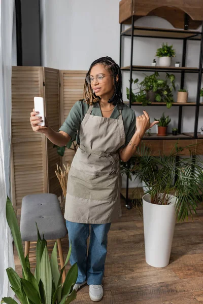 African american florist in apron pointing at assortment of green potted plants while having video call on smartphone — Stock Photo
