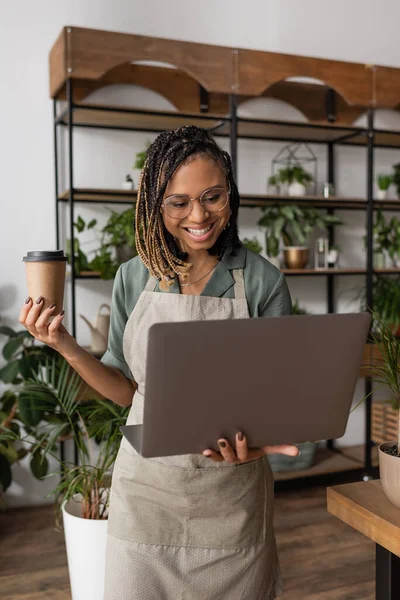 Florista americano africano com bebida takeaway sorrindo durante chamada de vídeo no laptop na loja de flores — Fotografia de Stock