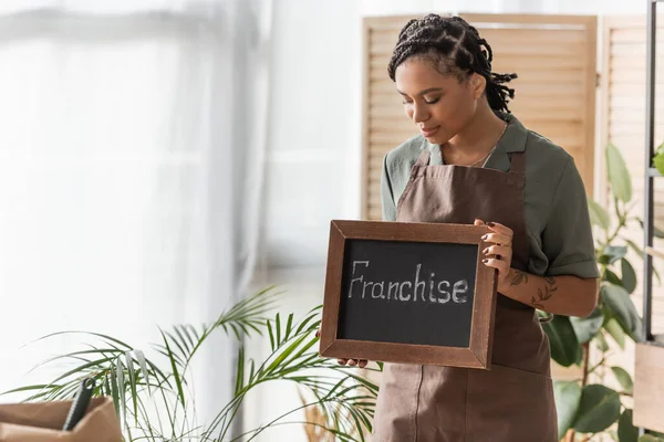 Pretty african american florist with trendy hairstyle holding board with franchise lettering in flower shop — Stock Photo