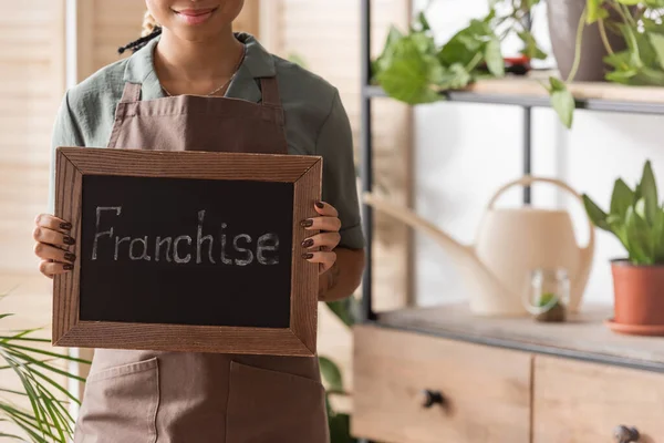 Partial view of happy african american florist in apron holding board with franchise inscription in blurred flower shop — Stock Photo