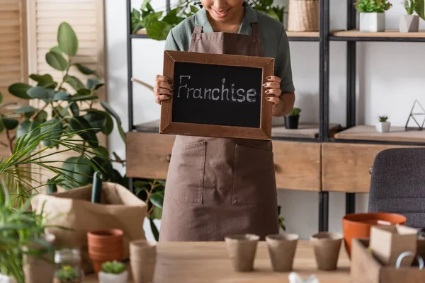 Cropped view of african american florist in apron holding board with franchise lettering near plants and flowerpots on blurred foreground — Stock Photo