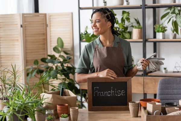 Pleased african american florist holding work gloves and board with franchise lettering while looking away in flower shop — Stock Photo