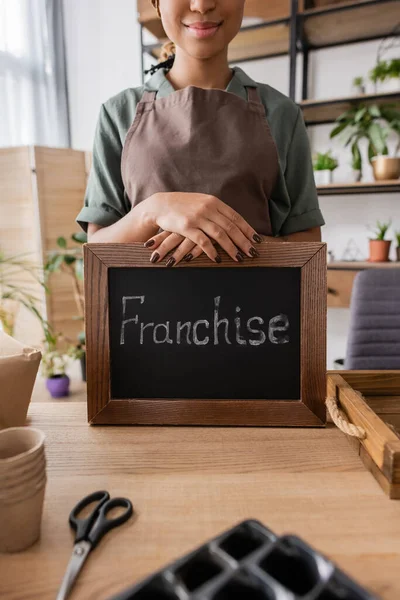 Partial view of african american florist holding board with franchise inscription near flowerpots and scissors on blurred foreground — Stock Photo