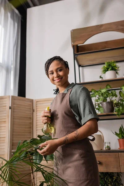 Young african american florist smiling at camera while standing with spray bottle near green plant — Stock Photo