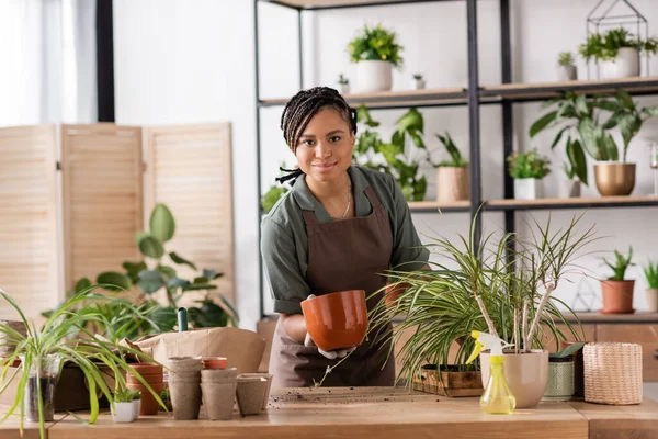 Happy african american florist holding flowerpot and looking at camera near plants and spray bottle on table — Stock Photo