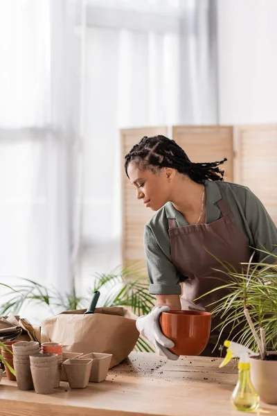 Stylish african american florist holding flowerpot and looking in paper bag with soil while working in shop — Stock Photo