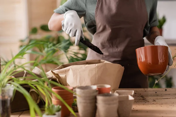 Cropped view of african american florist in apron holding flowerpot and garden scoop with soil near blurred plants — Stock Photo