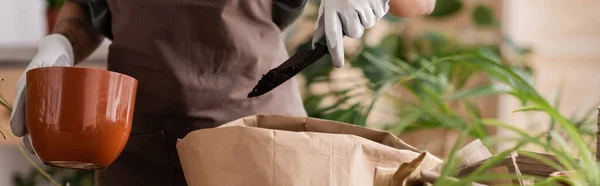 Обрезанный вид на африканского американского флориста, держащего цветочный горшок рядом с бумажным пакетом и садовым совок с почвой рядом с размытыми растениями, баннер — стоковое фото