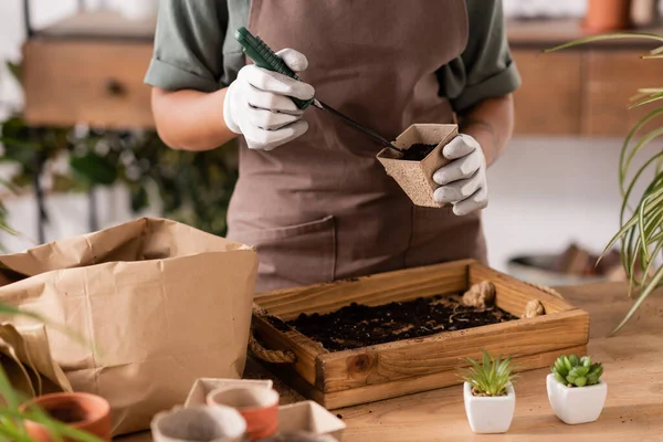 Cropped view of african american woman filling flowerpot with soil near wooden box and small plants — Stock Photo