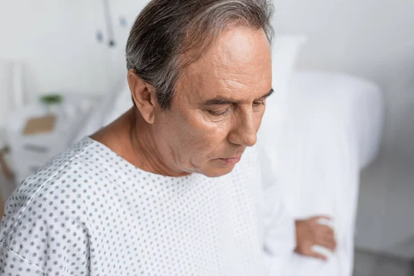 Sad senior man in patient gown looking down in hospital ward — Photo de stock