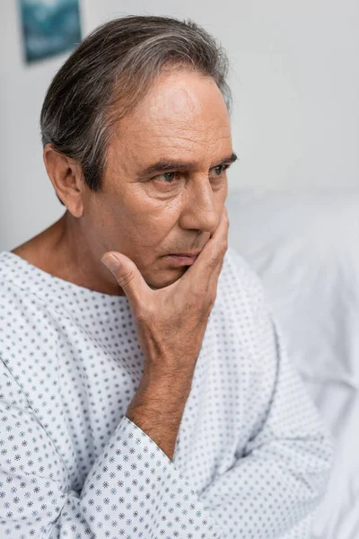Disappointed elderly man in patient gown looking away in hospital ward - foto de stock