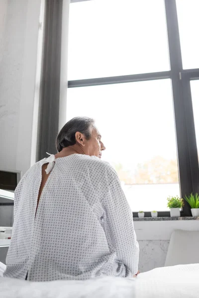Side view of senior patient in gown sitting on hospital bed — Stock Photo