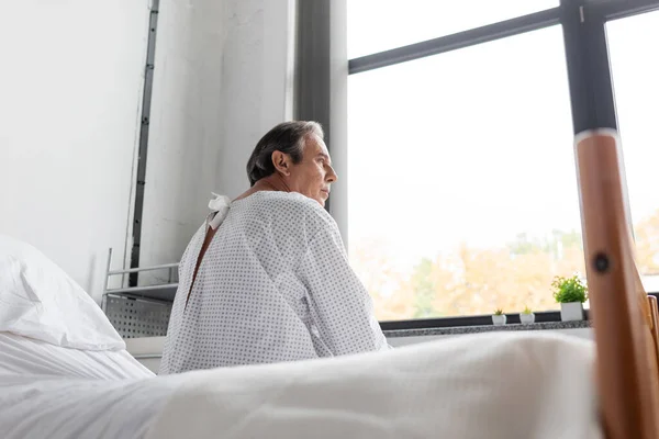 Side view of sick elderly man in patient gown sitting on blurred hospital bed — Stock Photo