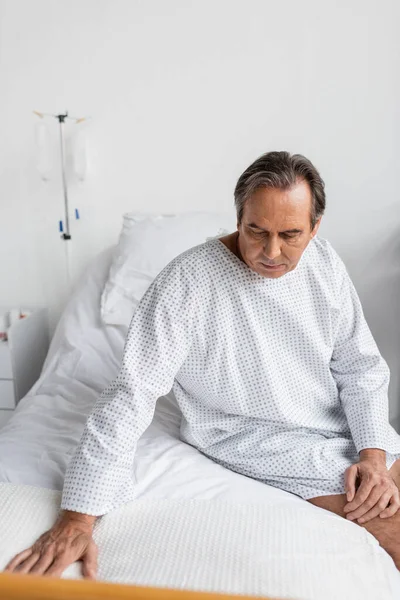 Upset senior patient in gown sitting on hospital bed — Stock Photo